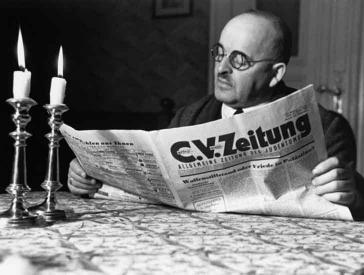 Schwarz-Weiß-Foto von Herbert Sonnenfeld: Ein Mann sitzt an einem Tisch mit zwei brennenden Schabbatkerzen und liest die C.V.-Zeitung
