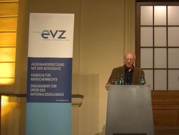 Brian Klug stands at a lectern and gives a lecture. To his left is a display with the logo of the Foundation “Remembrance, Responsibility, and Future” (evz)