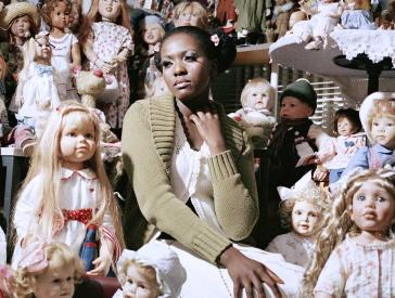 A young woman sits in a room filled with dolls.