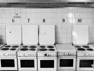 Seven consecutively numbered kitchen stoves are crowded together in a white-tiled kitchen (black and white photo).