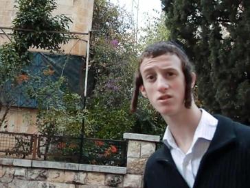 In a Jerusalem residential neighborhood, a young man with payes looks into the camera with a curiously surprised expression.