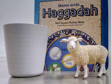 A toy sheep and a white container stand in front of a Haggadah for children. 