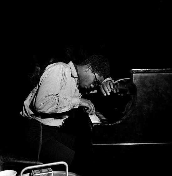 Black-and-white photo of Herbie Hancock in profile, he plays the piano with one hand, his other hand and his forehead lean on the piano