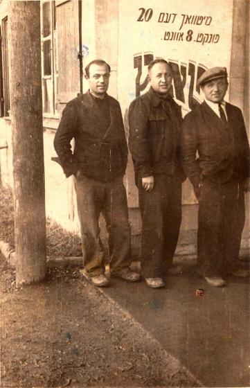 Black and white photo of three men, standing at a slight angle with their hands in their pockets 