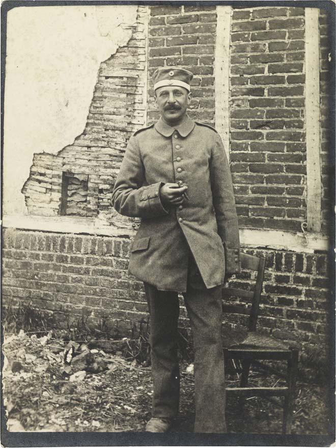 Black-and-white photograph: A soldier in uniform smoking in front of a building with cracked stucco.