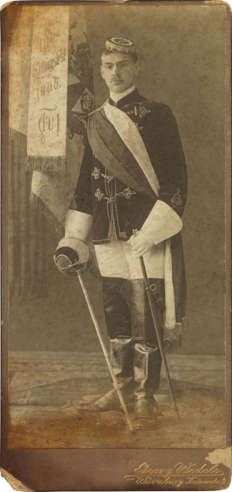Black-and-white photograph: student in full fraternity regalia with a fencing foil, fraternity flag in the background