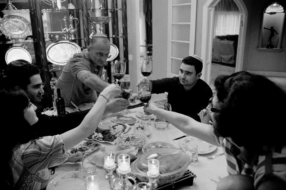 Black and white photograph of a group of people sitting at a dinner table with lots of food, they all raise their wine glasses in the middle of the table, the men are wearing kippahs