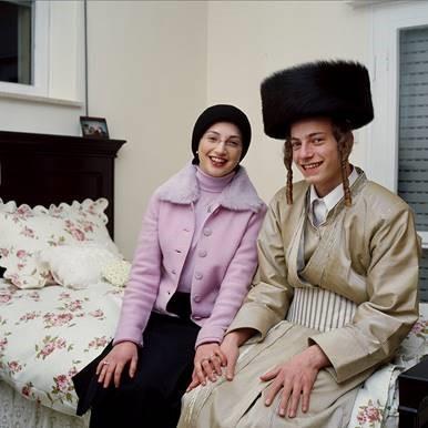 A woman with a headscarf and a man with temple curls and shtreimel sit on a bed laughing and holding hands.