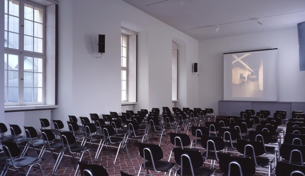 Rows of empty black chairs face the front of the room where a projector screen is located