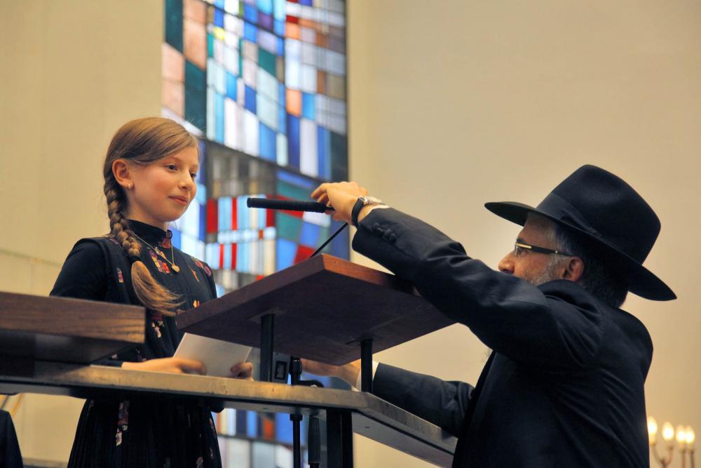 A young girl at a lectern, a man in orthodox clothing adjusting the microphone