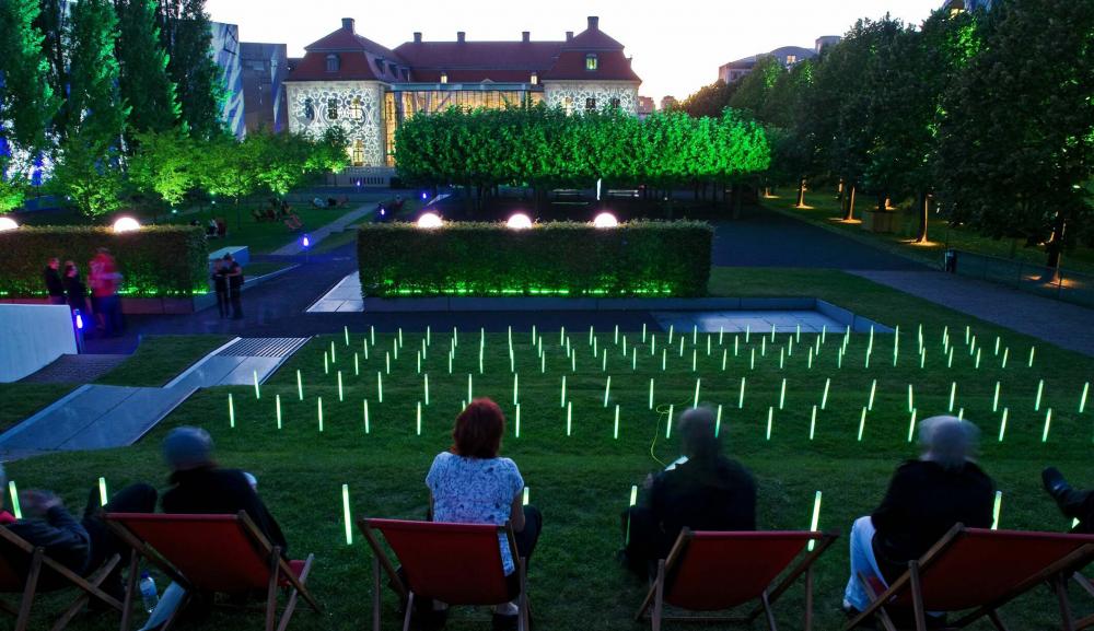 People sit in red lawn chairs in the Museum Garden, the garden is lit up brightly under the darkening sky