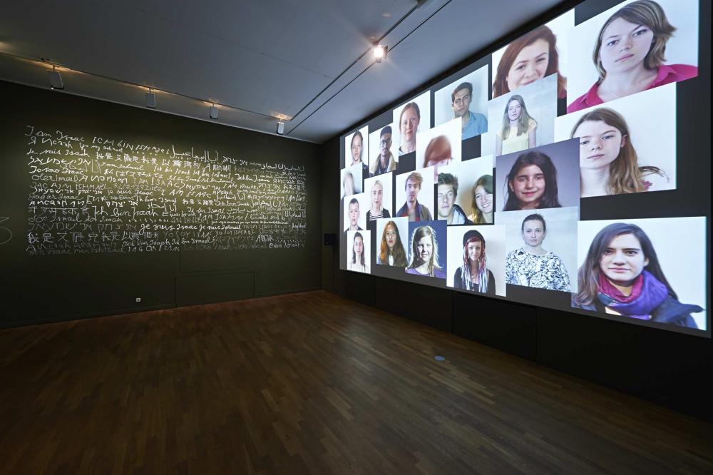 An empty room facing two walls, one wall is covered in portrait photos, the other wall is covered in text of various languages