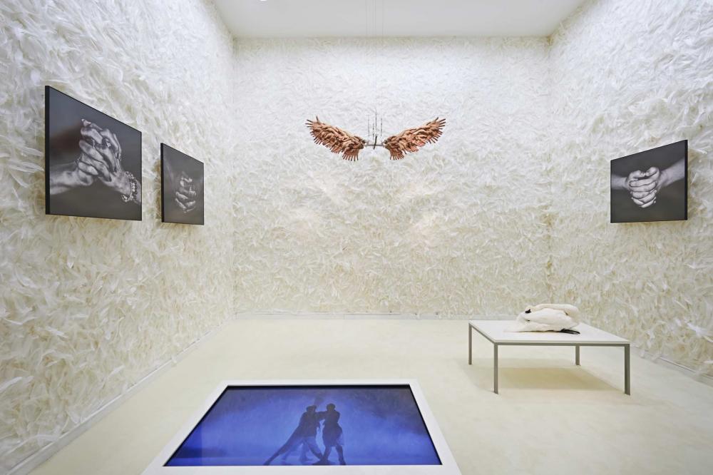 Mit Federn tapezierter Raum, an den Wänden Fotos gefalteter Hände, an der Decke hängt eine Skulptur aus Händen, die ein Flügelpaar bilden