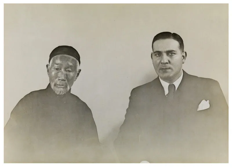 Black and white picture. On the left an older man in Chinese clothing, and on the right a middle-aged man in a suit. Both are looking into the camera.