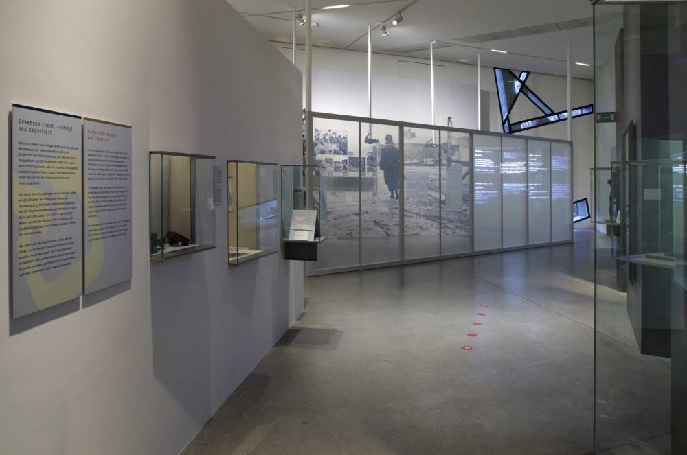 Ausstellungsraum: links Textafeln und Wandvitrinen, im Hintergrund eine halbhohe Wand, auf die Fotos und Text projiziert werden