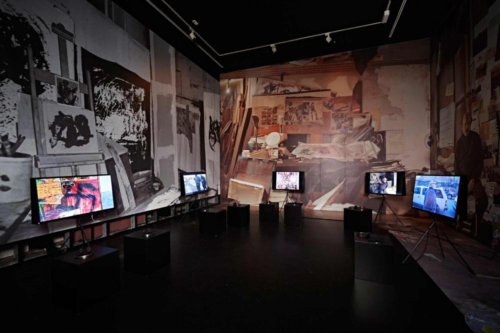 Exhibition space, the walls are covered with large photographs of Boris Lurie's studio, TV screens fill the room