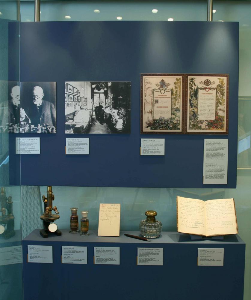 Historische wissenschaftliche Objekte wie ein Mikroskop und Glas, Flüssigkeitsflaschen und handschriftliche Notizen werden unter Bildern und Texten gezeigt.