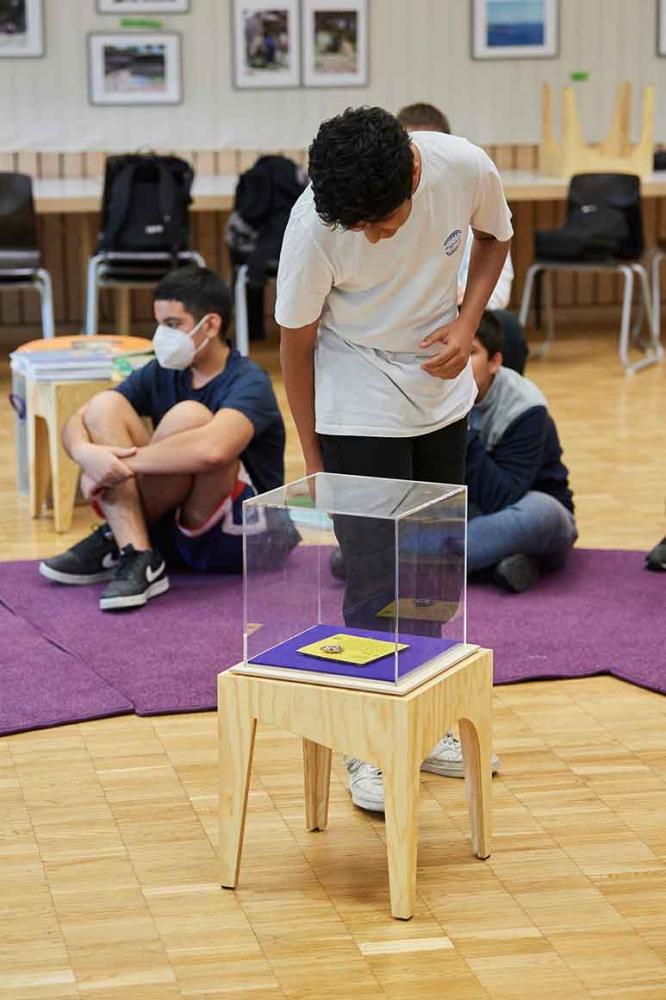 On a wooden stool is a display case containing a yellow card on a purple background, which a young person is looking at.