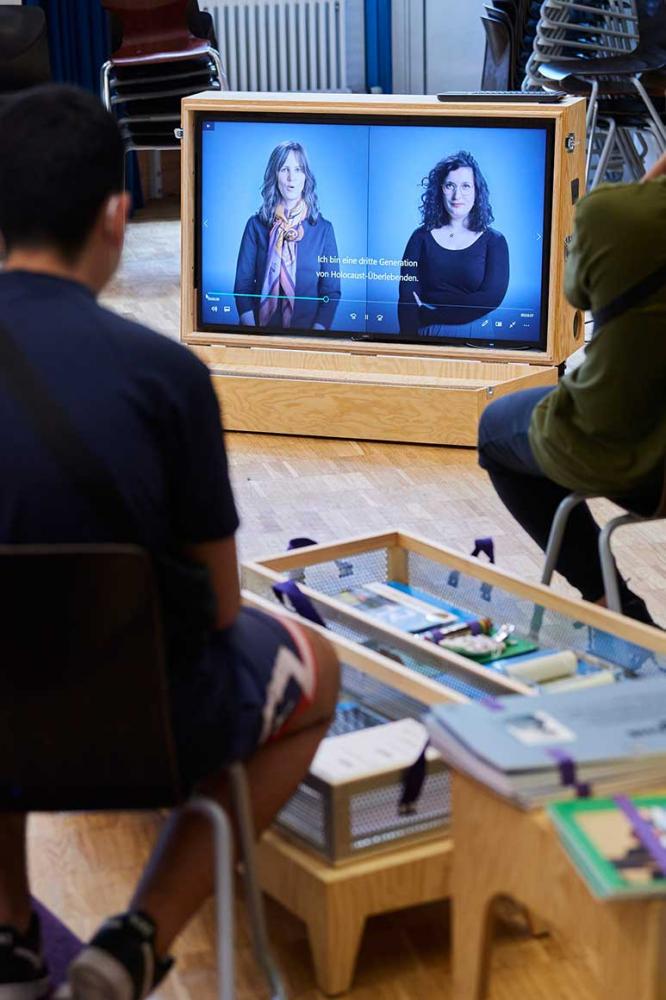Jugendliche sitzen vor einem Bildschirm, auf dem zwei Frauen nebeneinander zu sehen sind.