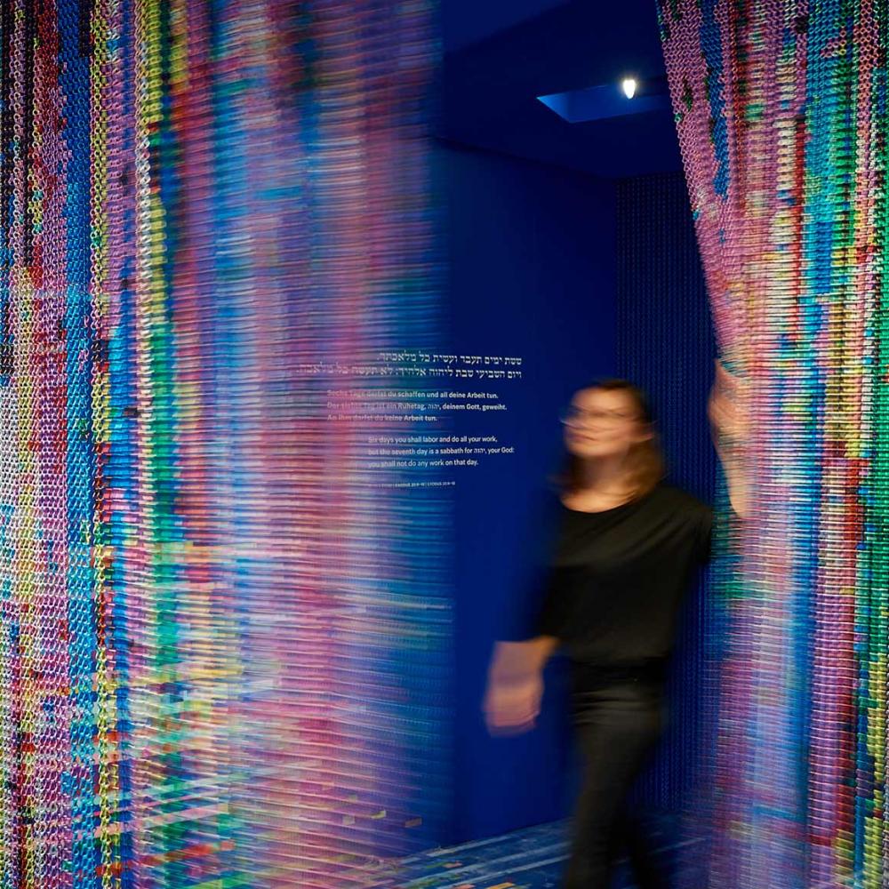 A person walks through a colourful glittering curtain, on the wall the commandment to rest on the seventh day (in Hebrew, German and English)