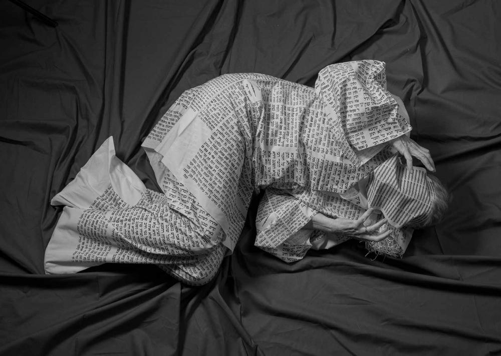 A person wrapped in paper printed with Hebrew letters lies on a dark, wrinkled sheet.