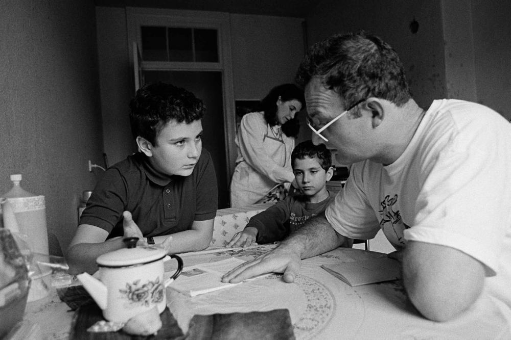 Black and white photo of a man wearing glasses talking to a young boy at a small kitchen table, the man points to a book at the table and the young boy raises his hand as if to ask a question