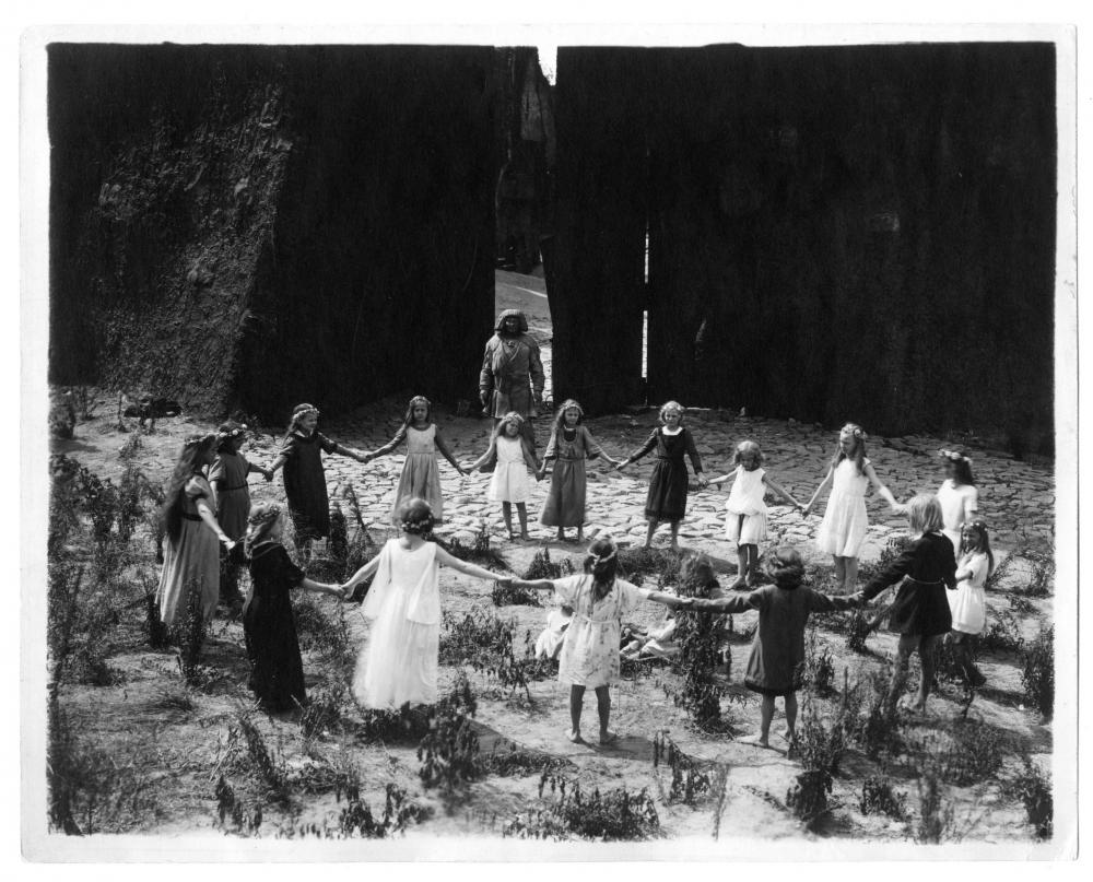 Filmstill from the movie “The Golem, How He Came Into the World” by Paul Wegener: children are holding hands and dancing in a circle, behind them the golem stands watching.