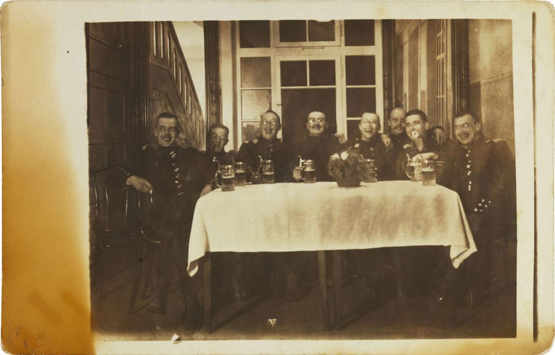 Black-and-white photograph: eight soldiers in uniform sitting around a table with beer steins and laughing, a window can be seen in the background