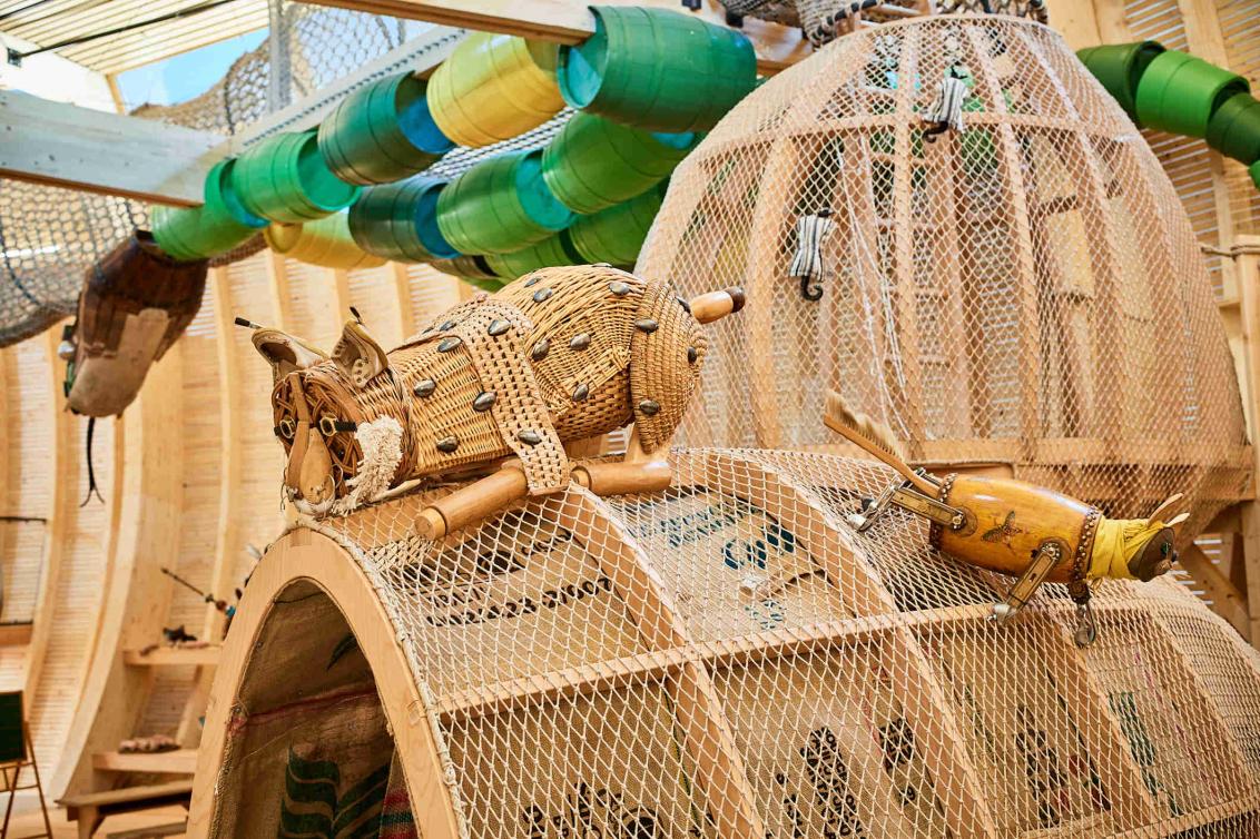 A lurking lynx made of everyday objects sits on a hut made of wood and net. In the background, a snake is meandering on the ceiling. It is composed of barrels