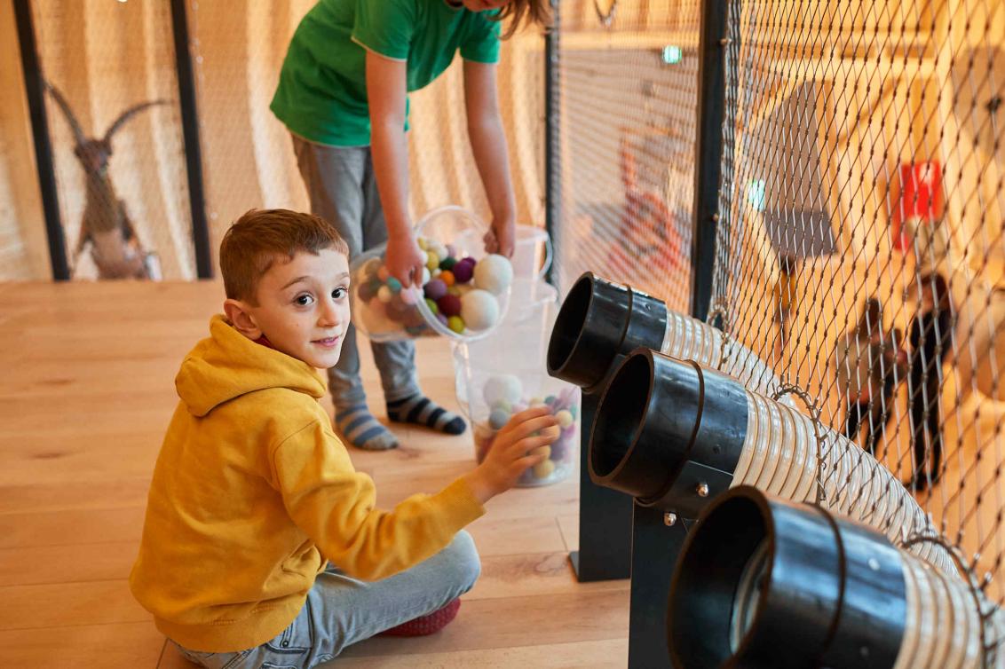 Child wearing a yellow sweatshirt is sitting in front of pipe openings into which he has thrown something. In the background another child filling balls from one transparent bucket into another