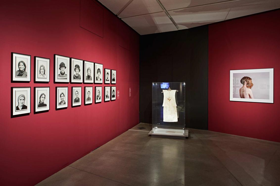 View of a room in the exhibition Cherchez la femme with portraits of women on one wall, a dress in a glass case, and a photo of a woman with two wigs on another wall