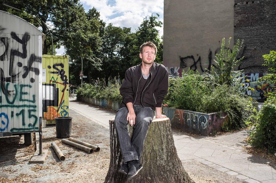 Foto: Ein Mann, vor Bäumen und mit Graffiti besprühten Containern auf einem Baumstamm sitzend