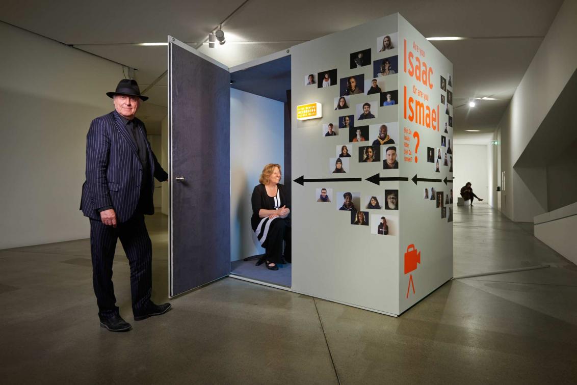 A man hold the door open to a small isolated room with a smiling woman sitting inside, the outisde of the room is covered in portrait photos