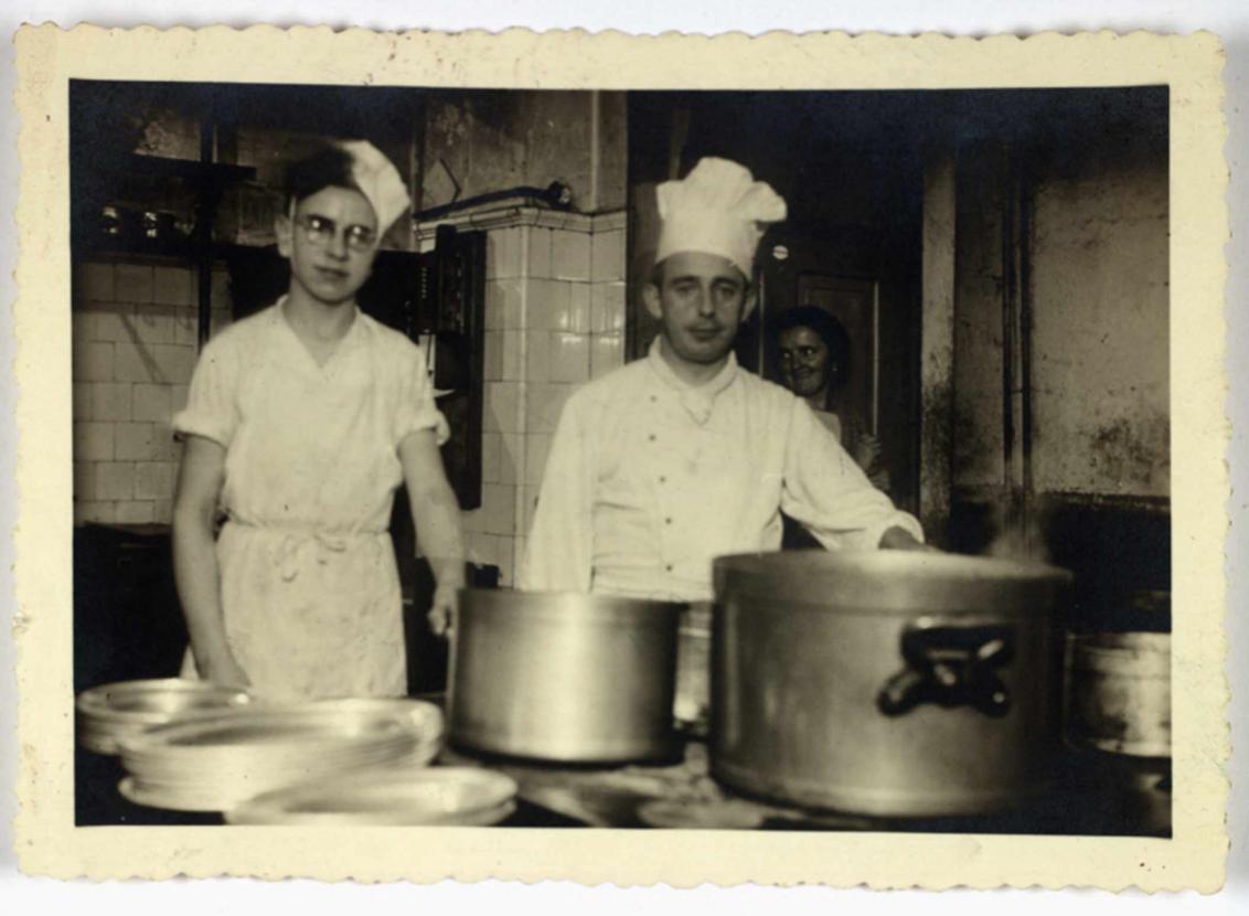 Henry Wuga mit Kochlehrlingsmütze in der Hotelküche, neben ihm ein Koch, im Hintergrund eine weibliche Küchenhilfe.