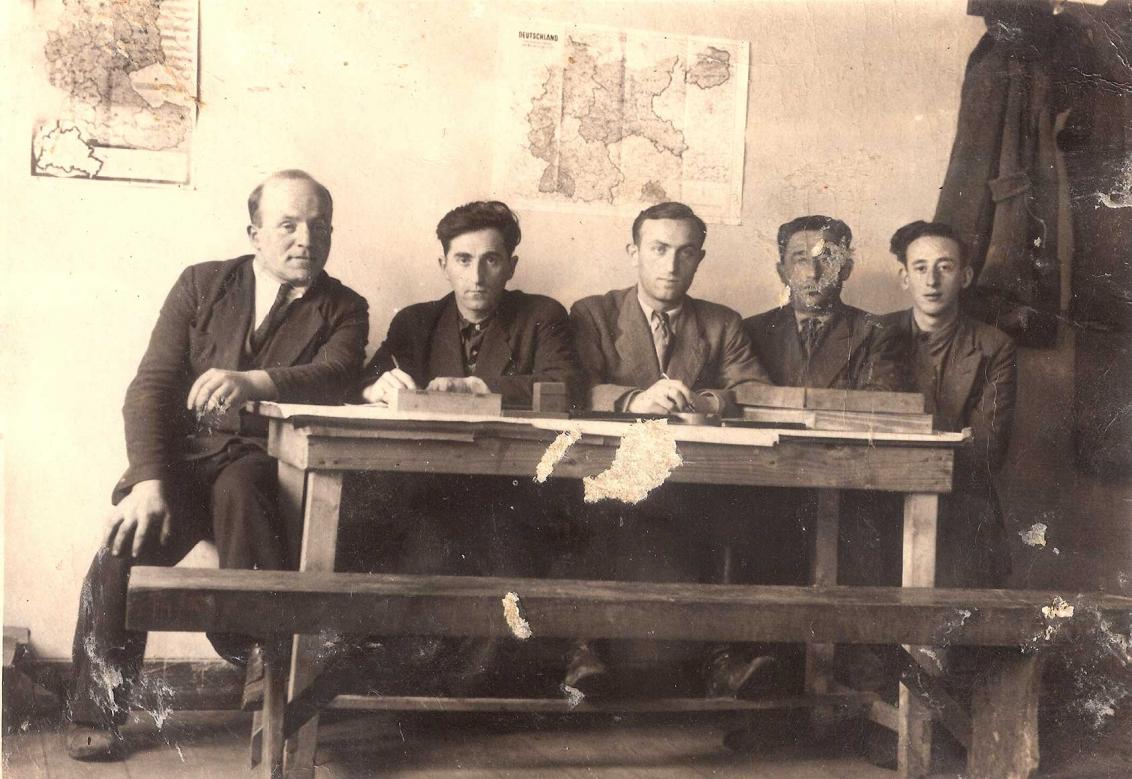 Black and white photograph of five men sitting side by side at a wooden table with two maps hanging on the wall behind them