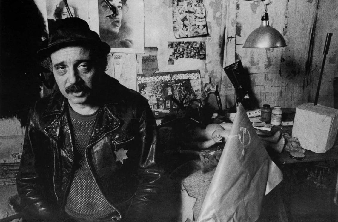 Black and white photograph of Boris Lurie in his studio, the walls and desk are filled with pictures and projects, Lurie wears a leather jacket with the Star of David