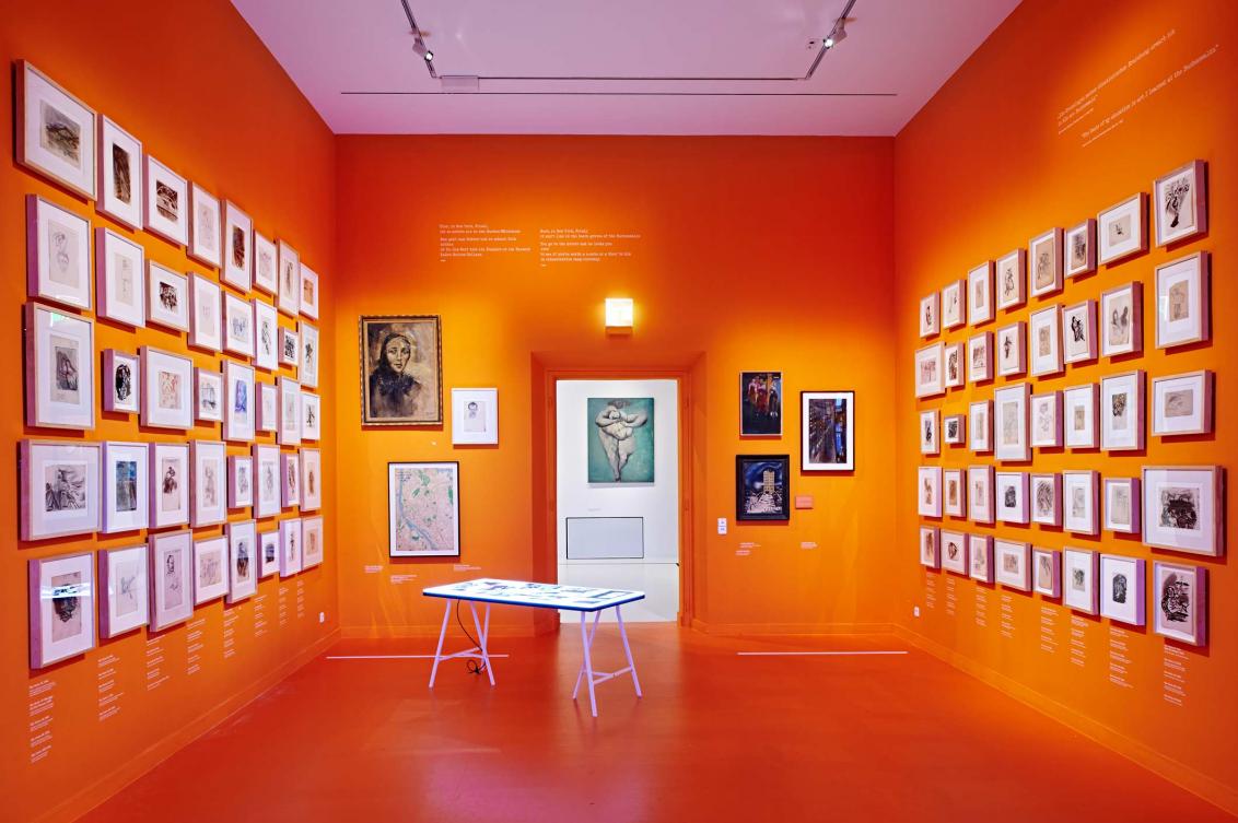 Ausstellungsraum mit orangen Wänden, an denen viele gerahmte Zeichnungen und Ölbilder hängen