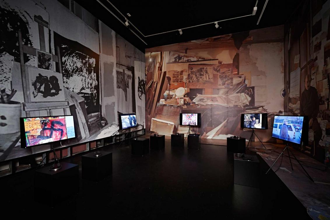 Ein Ausstellungsraum mit mehreren Bildschirmen, an den Wänden ist Fototapete angebracht, die die Wände von Boris Luries Atelier zeigt