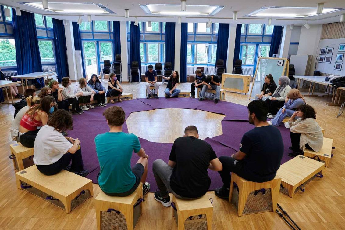 Young people sit on boxes made of wood and metal around a purple circular carpet.