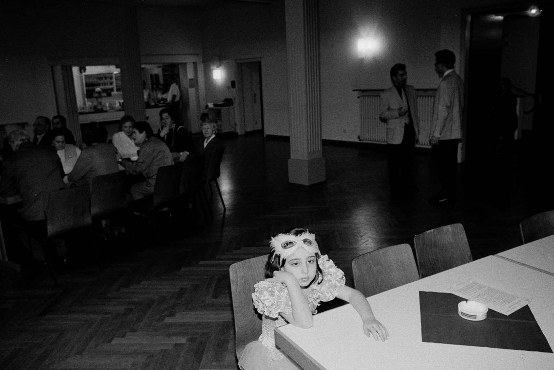 Ein verkleidetes Mädchen sitzt allein an einem Tisch in einem Festsaal, im Hintergrund Erwachsene in Gruppen im Gespräch (Schwarz-Weiß-Foto)
