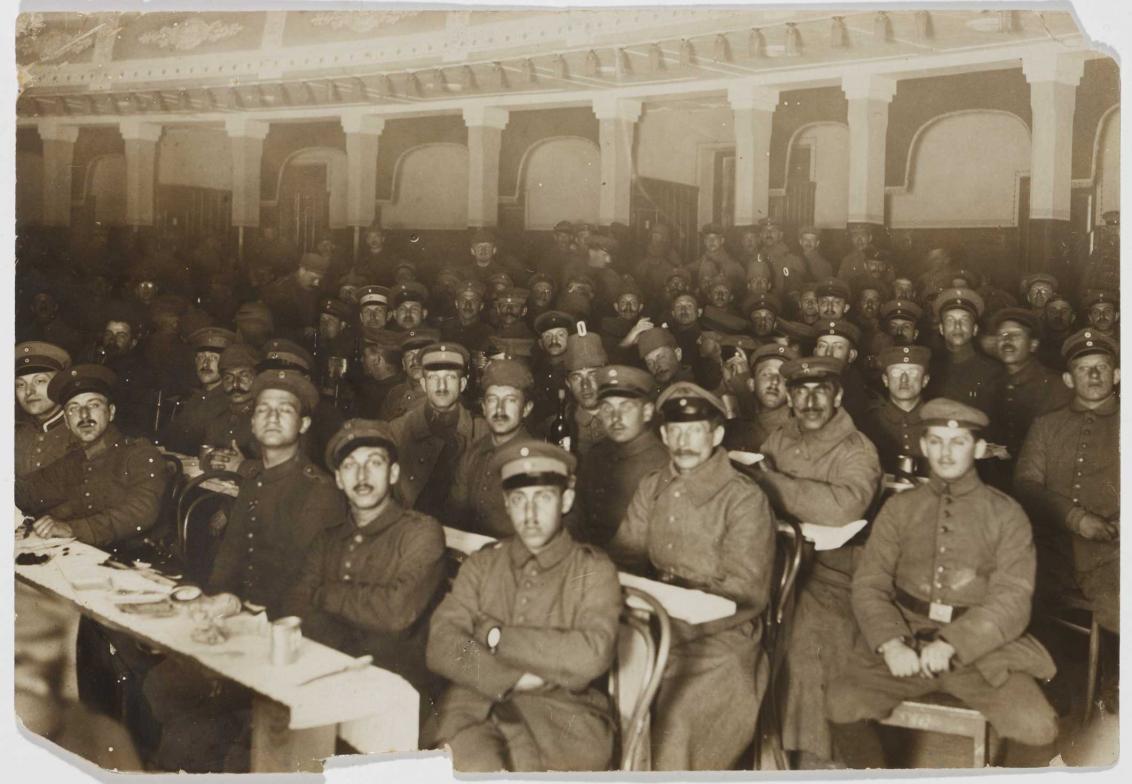 Soldaten sitzen uniformiert mit bedeckten Köpfen an langen Reihen improvisierter Tische; der vorderste ist gedeckt. Die Aufnahme wurde von einem Standpunkt rechts neben den Bankreihen aufgenommen
