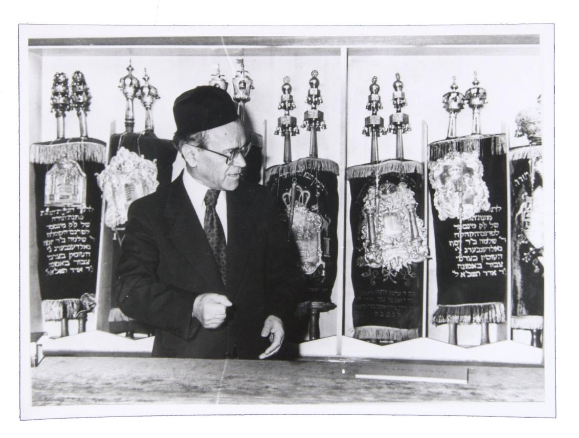 Black and white photo of man in suit in front of nine Torah scrolls