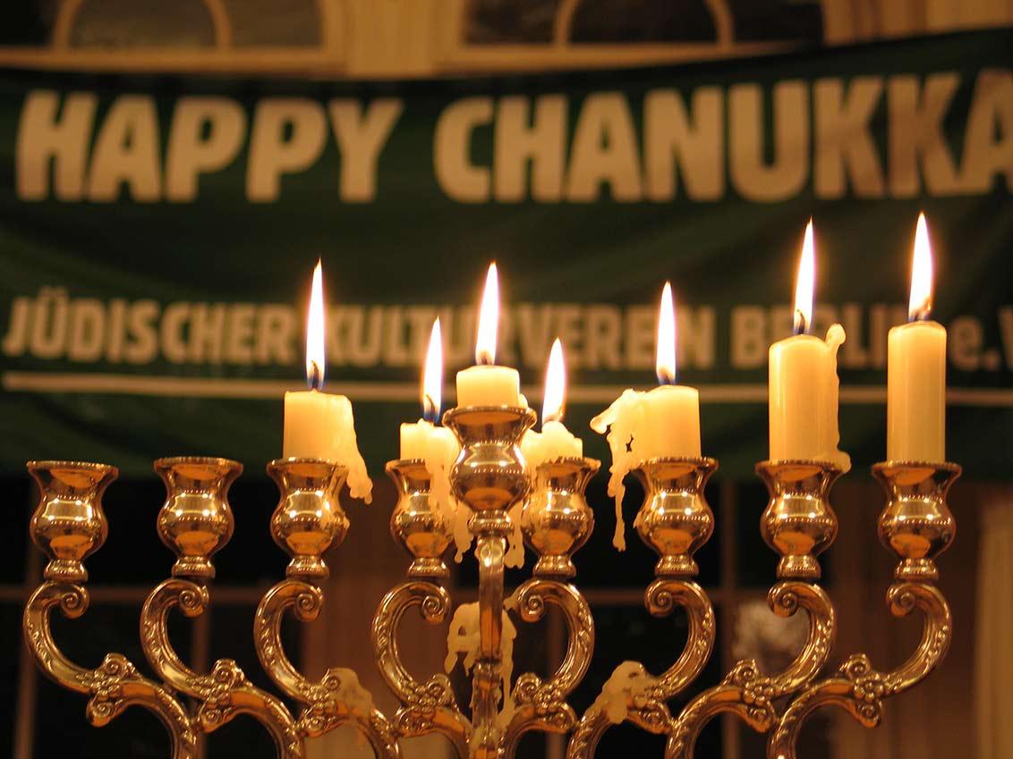 Chanukkaleuchter mit brennenden Kerzen, dahinter ein Banner mit der Aufschrift: Happy Chanukka. Jüdischer Kulturverein Berlin.