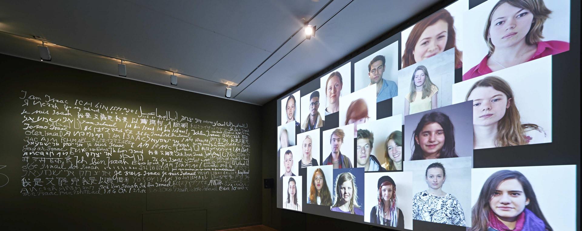 A wall reading »I am Isaak/Ismail« in several languages to the right and projected images of young peoples' faces to the lefte left 