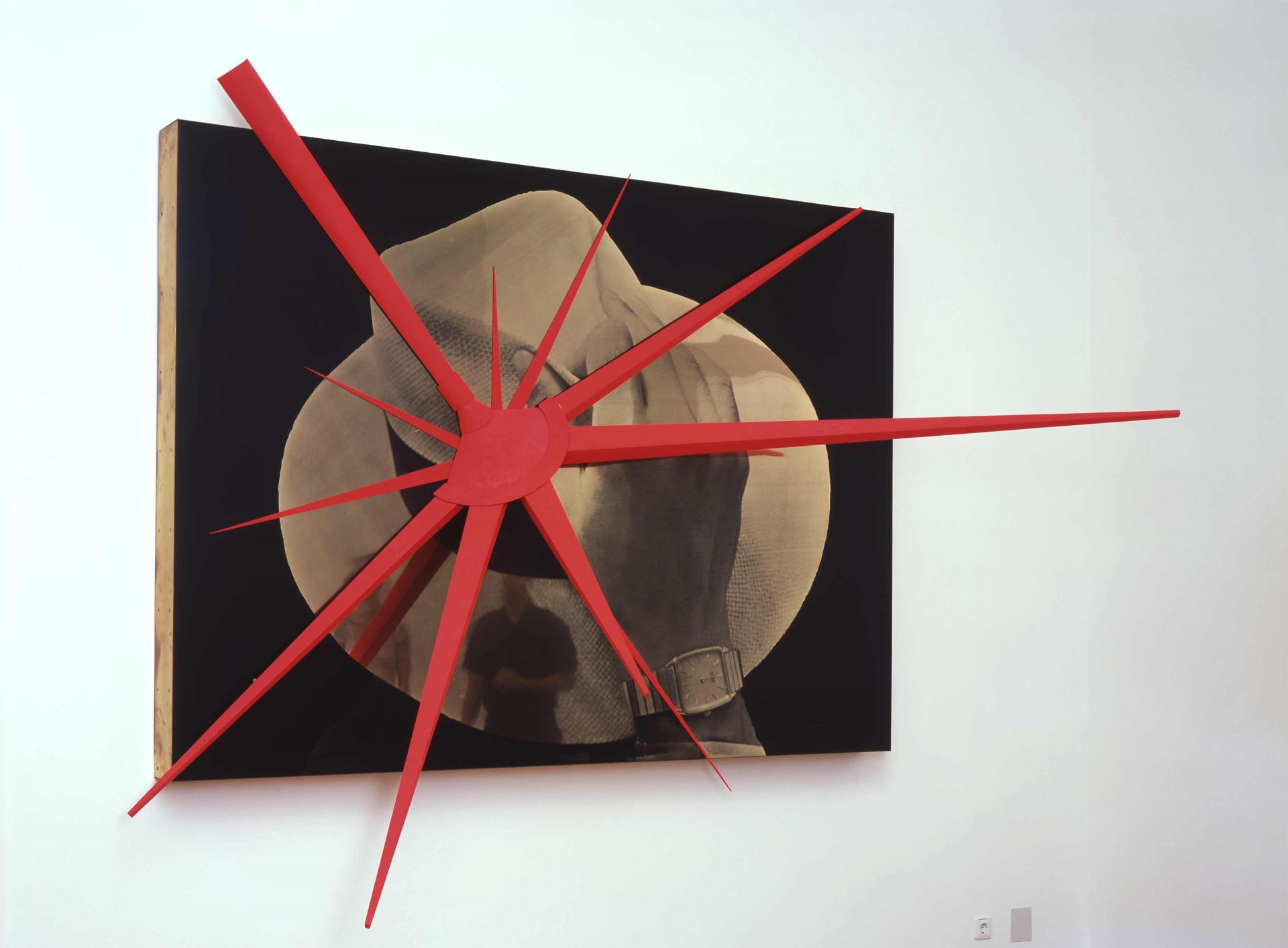 The large-scale artwork shows a highly magnified photo of a hat and a hand about to lift the hat. On the photo is mounted a red three-dimensional virus-like object