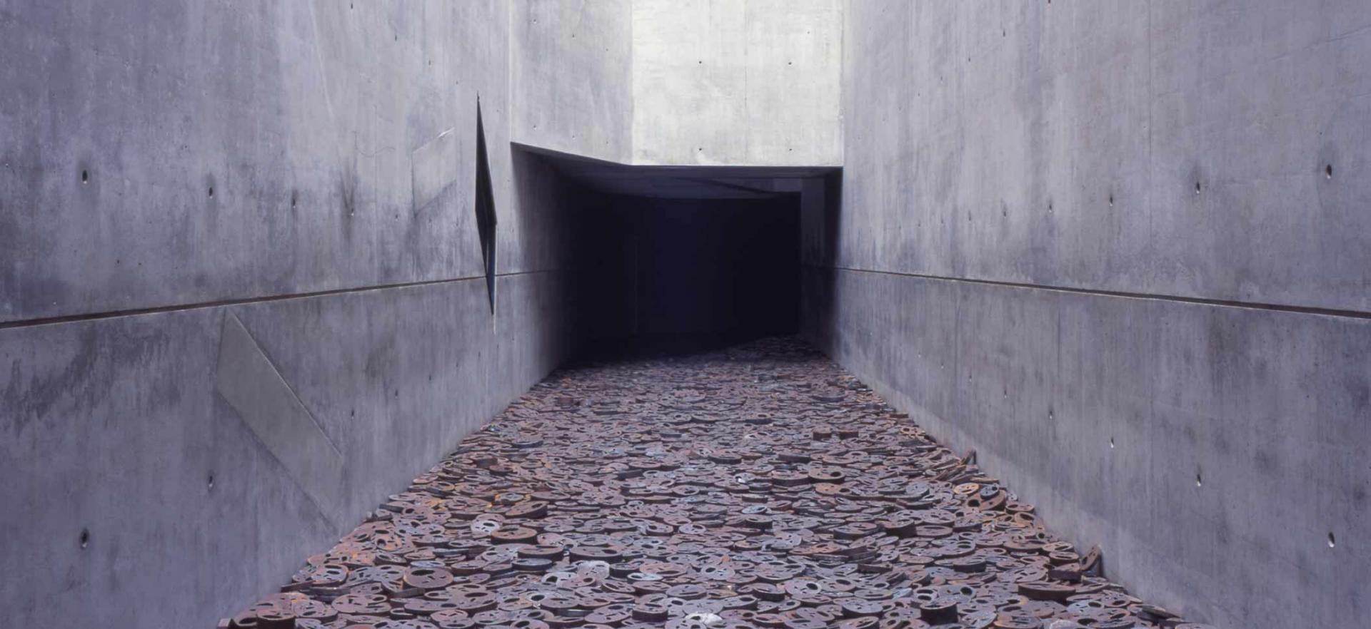A corridor with bare concrete walls that gets lost in the dark. The floor is littered with thick metal discs into which faces with torn open mouths have been sawn.