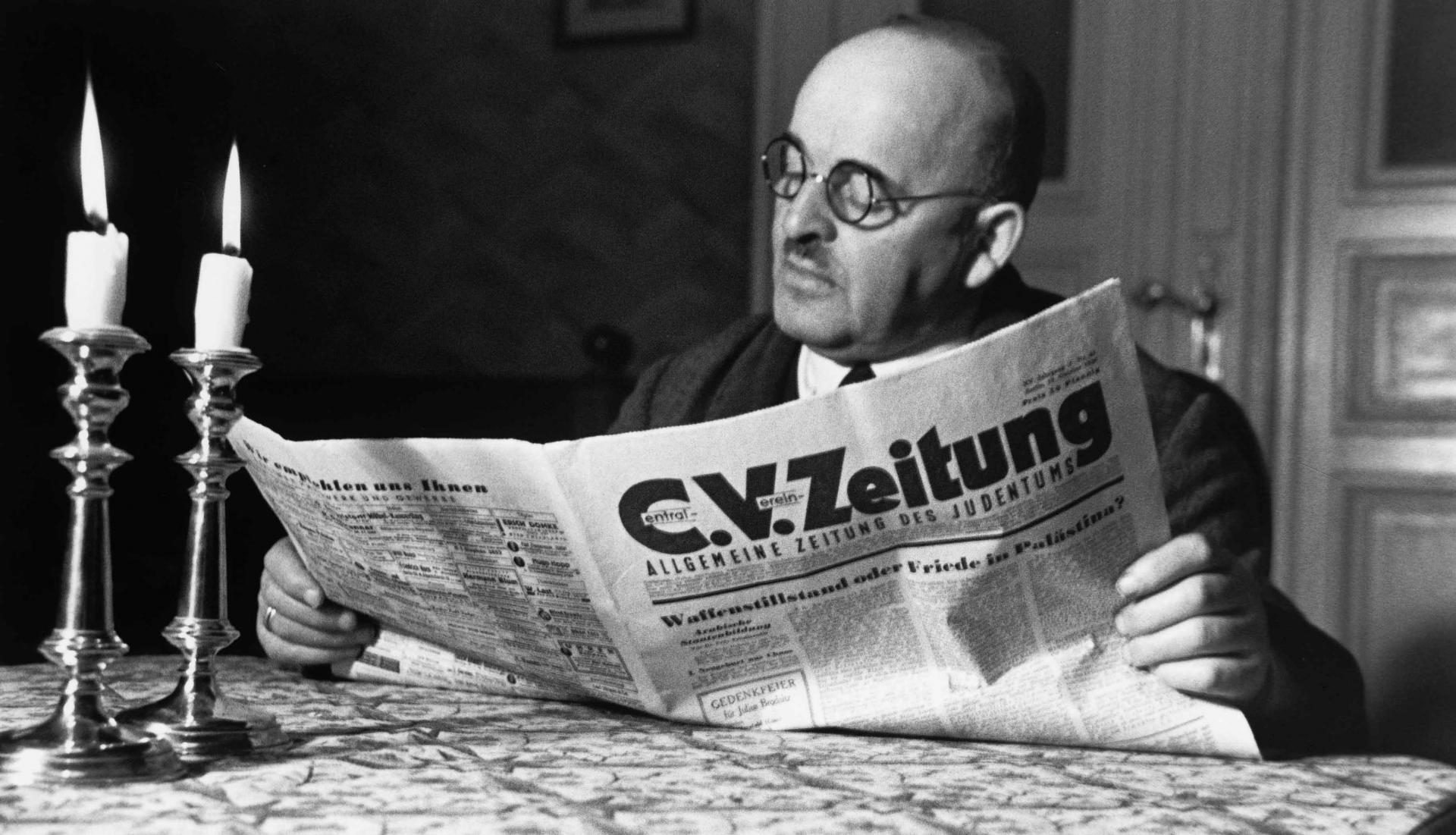 Schwarz-Weiß-Foto von Herbert Sonnenfeld: Ein Mann sitzt an einem Tisch mit zwei brennenden Schabbatkerzen und liest die C.V.-Zeitung