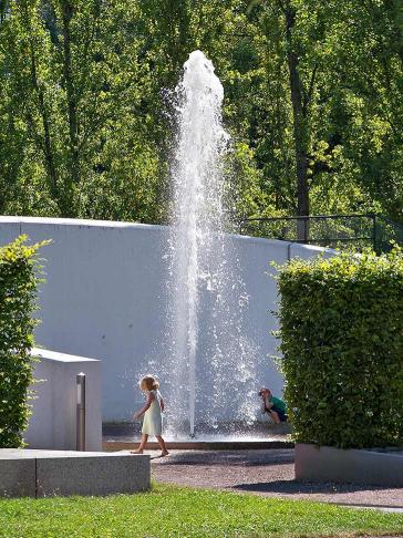 Una fuente de agua en un jardín, con un niño sentado a su lado, en primer plano una chica con un vestido de verano