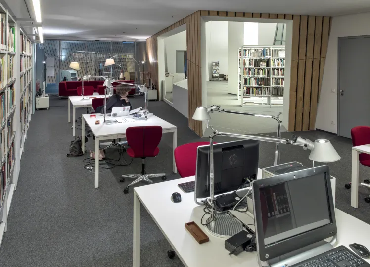 A row of desks, some equipped with desktops, stand in a room flanked by bookshelves. A passageway opens to the library area.