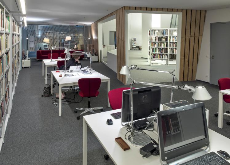 A row of desks, some equipped with desktops, stand in a room flanked by bookshelves. A passageway opens to the library area. 